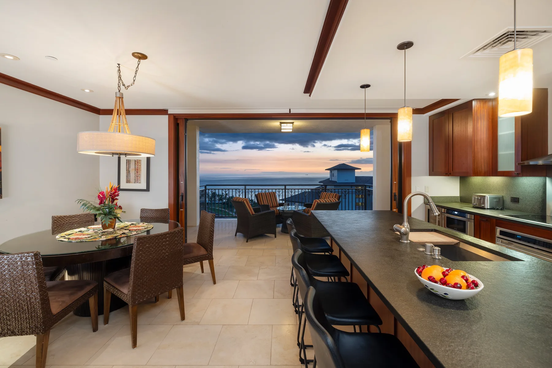 An open dining and kitchen area overlooks a balcony and the sunset sky.