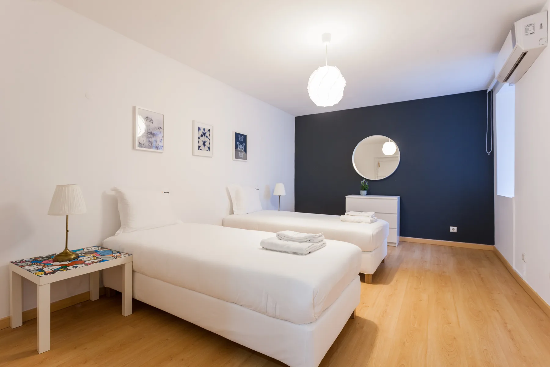 Un dormitorio luminoso con ropa de cama blanca, una pared oscura y un espejo redondo.