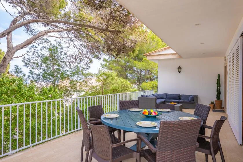 A covered outdoor dining area has a table and seating overlooking leafy trees.