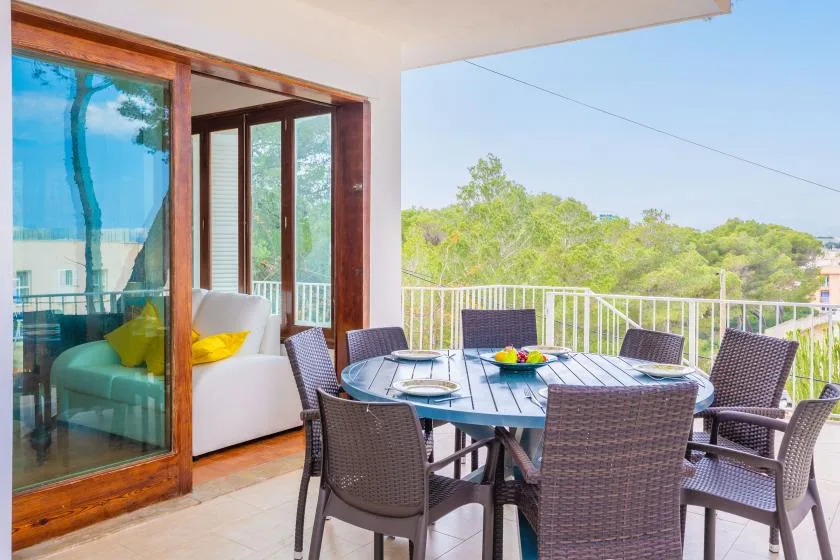 A furnished outdoor dining area sits beside large glass doors overlooking green trees.