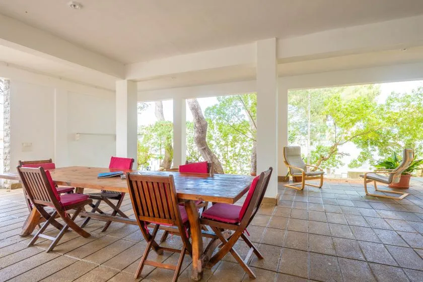 A covered dining area with a wooden table and chairs beside large open windows.