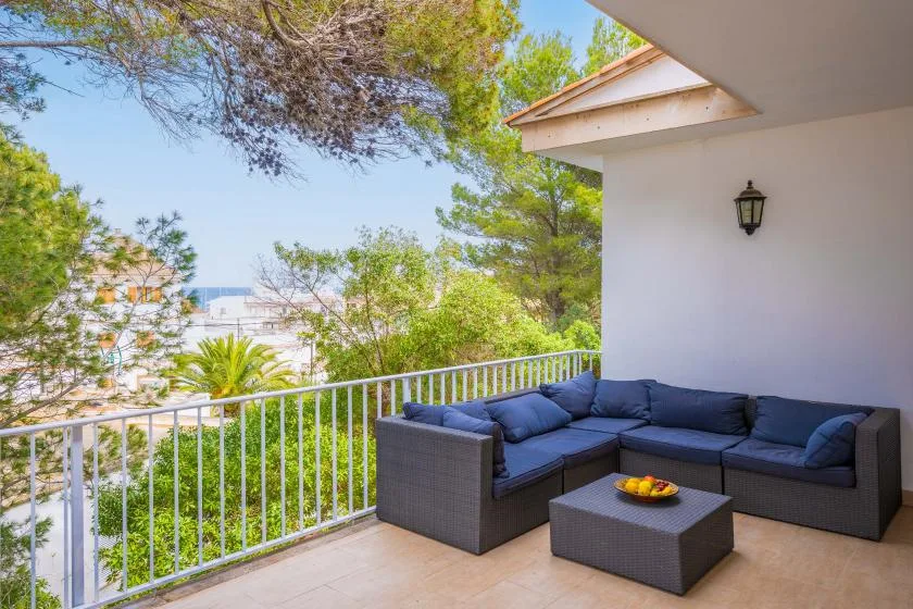 A balcony with outdoor seating overlooks leafy trees and a glimpse of the sea.