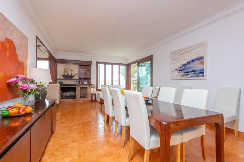 A dining area with a long table and white seating beside bright windows.