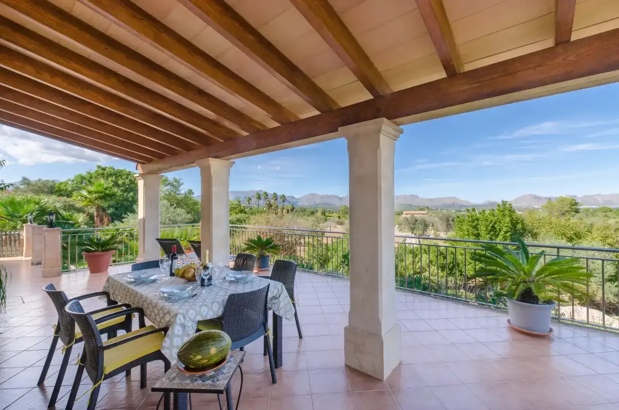 A covered outdoor terrace with a dining table and views of greenery and hills.