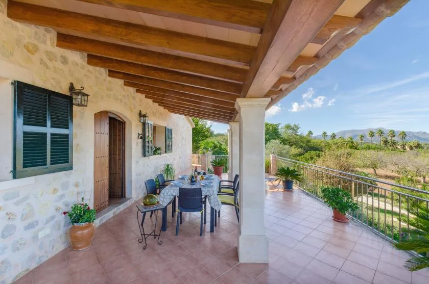 A covered outdoor terrace with a dining table and chairs beside a stone house.