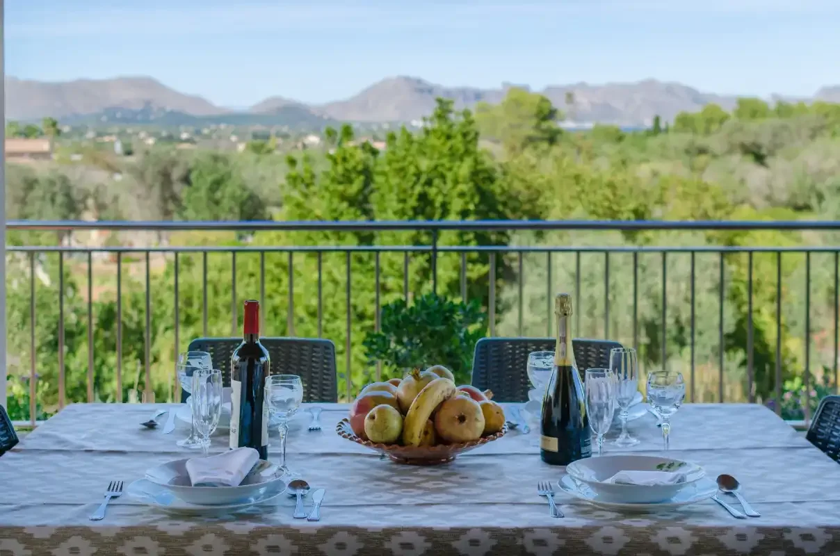 An outdoor dining table with fruit and place settings overlooks a green landscape.