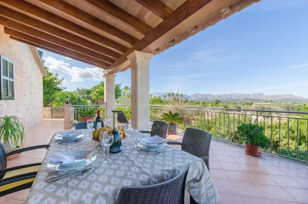 A covered terrace has a dining table overlooking greenery and distant hills.