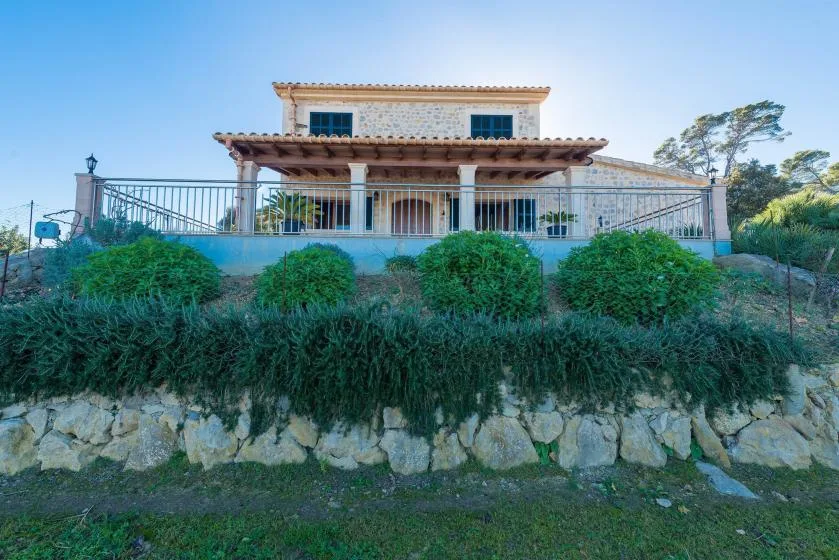 A two-story house with a broad covered terrace stands above a stone retaining wall.