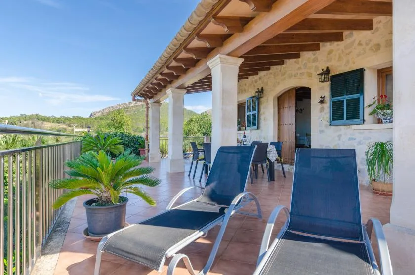 A covered stone terrace with lounge seating, potted plants, and views of green hills.