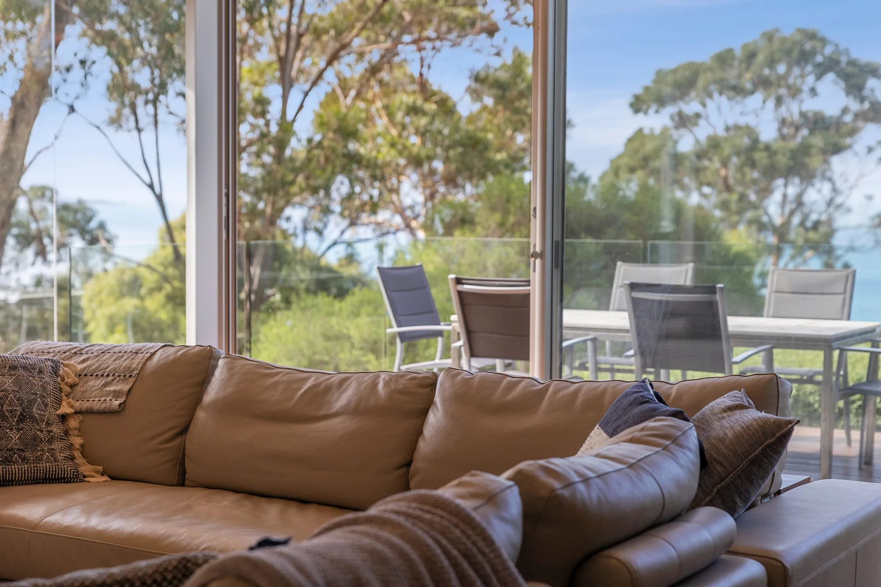 Una sala de estar con asientos junto a grandes ventanas que dan a árboles y una mesa exterior.