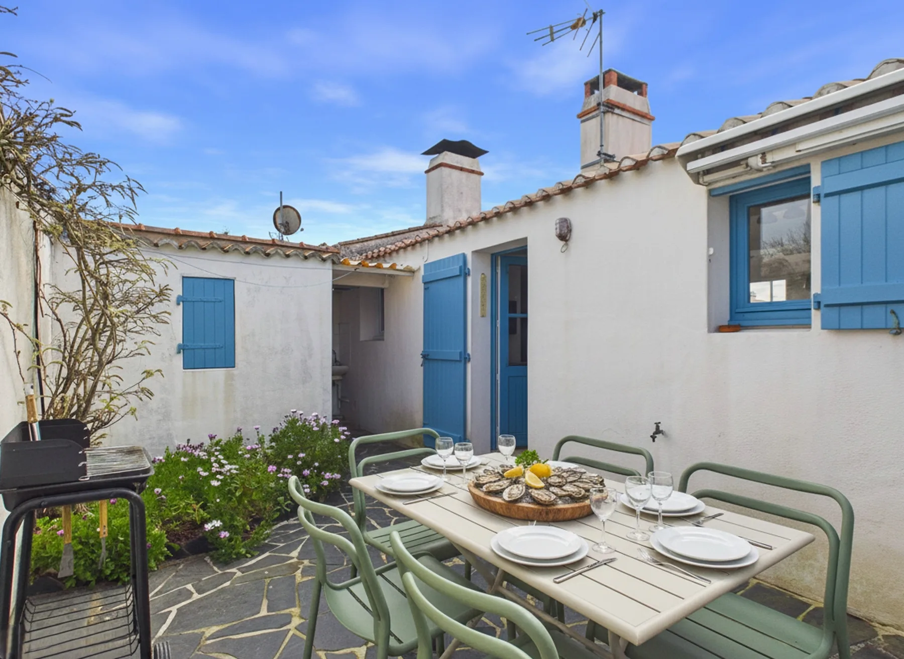 An outdoor dining area sits beside a white house with blue shutters.