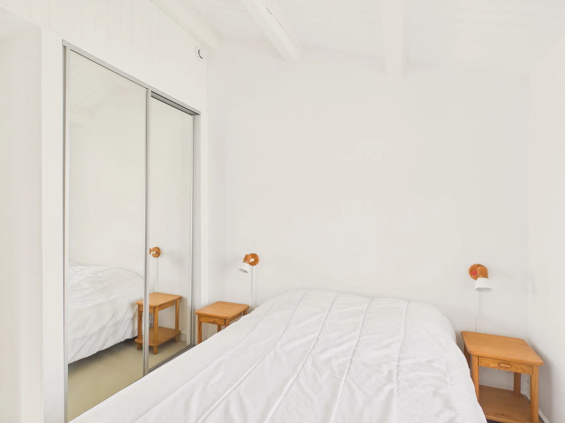 A bright bedroom with a bed, mirrored closet doors, and wooden bedside tables.