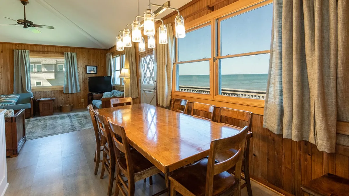 A wood-paneled dining area has a long table beside large windows overlooking the water.