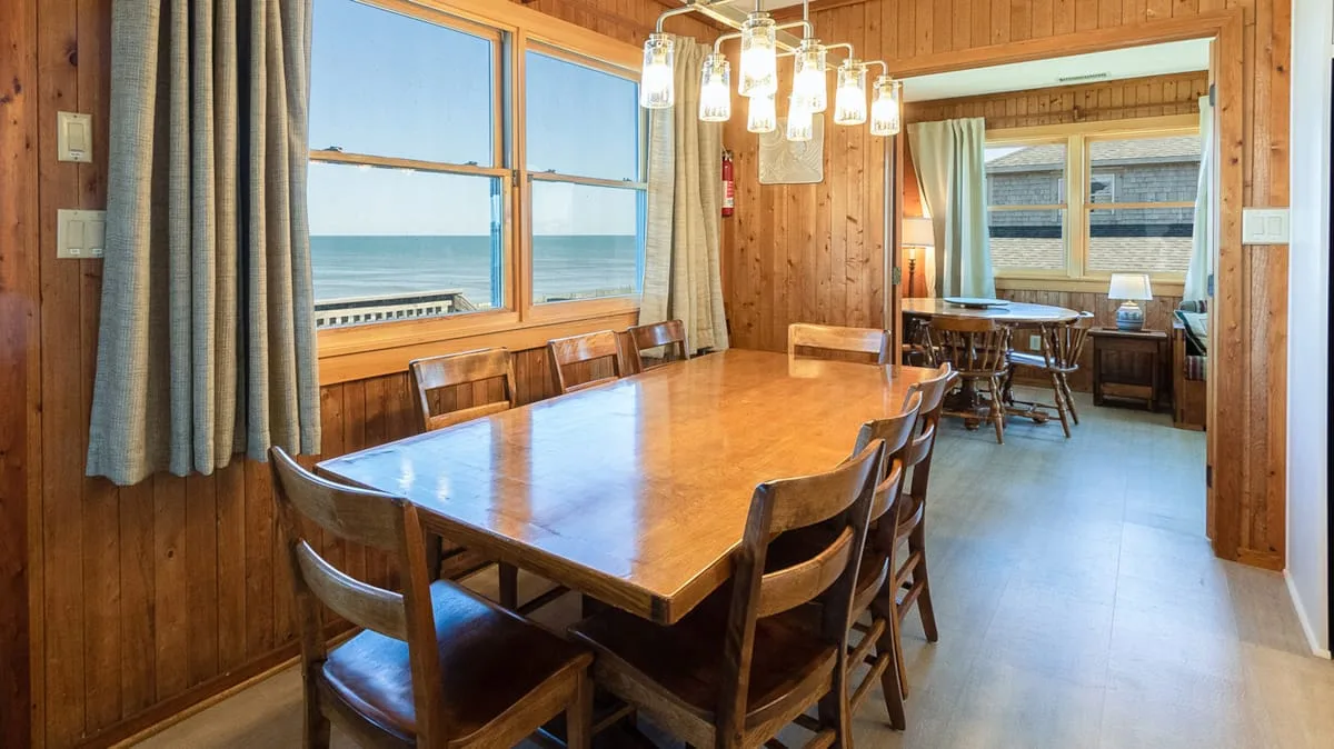 A wooden dining area with a long table beside large windows overlooking the water.