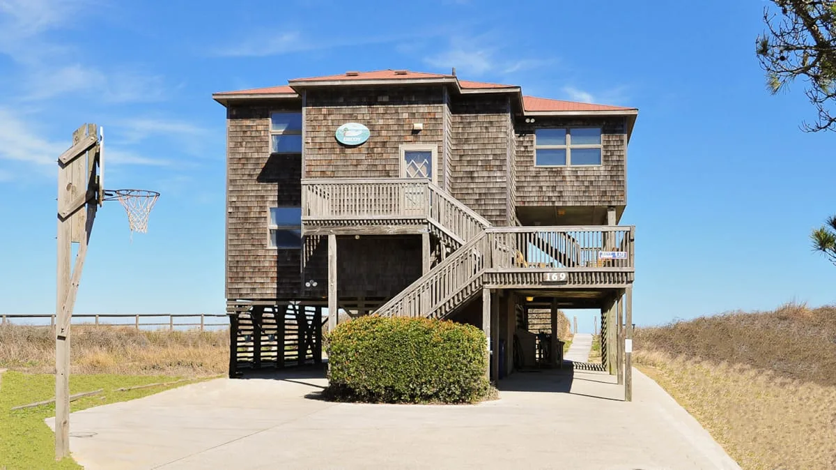 A raised coastal house stands beside a paved driveway and basketball hoop.