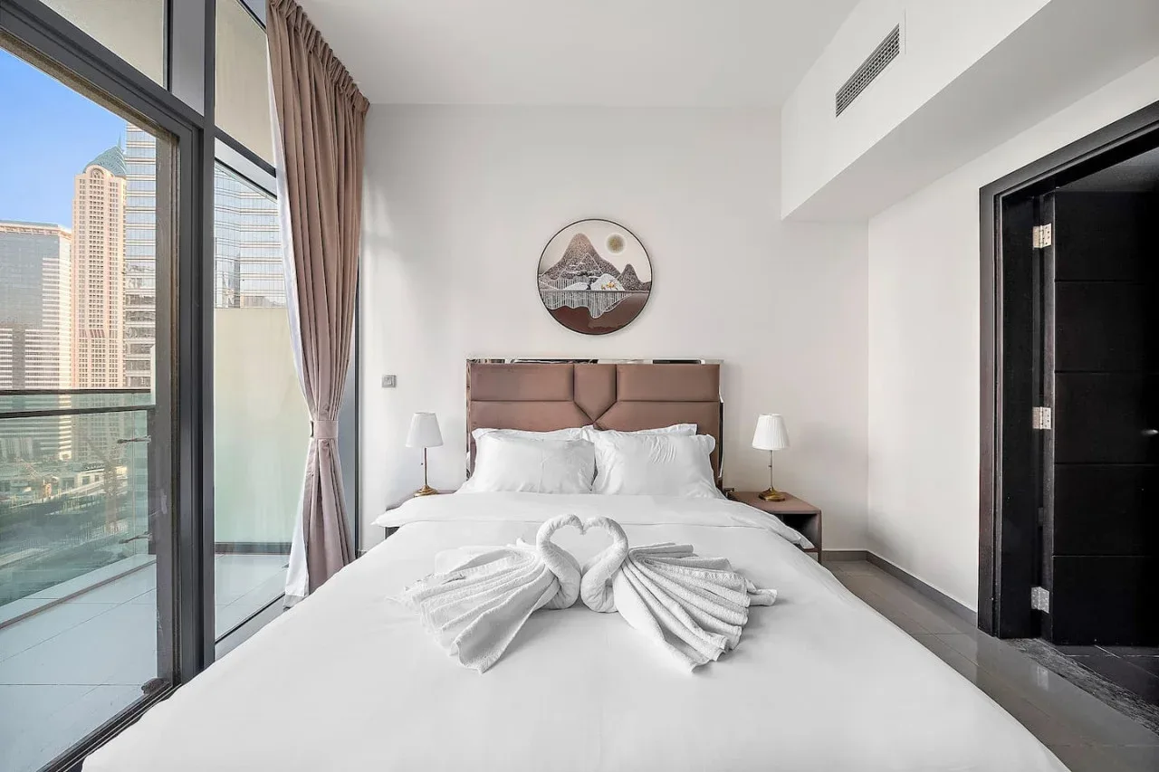 A bedroom with a neatly made bed, bedside lamps, and large windows.