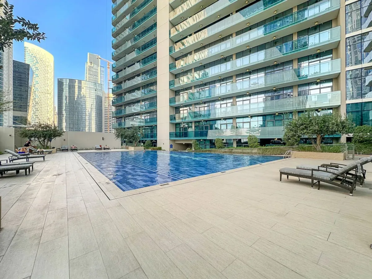 An outdoor swimming pool and lounge seating sit beside a modern high-rise building.