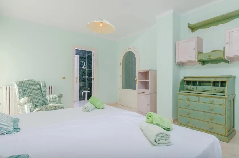A pastel bedroom with a bed, a patterned armchair, and a pale green dresser.