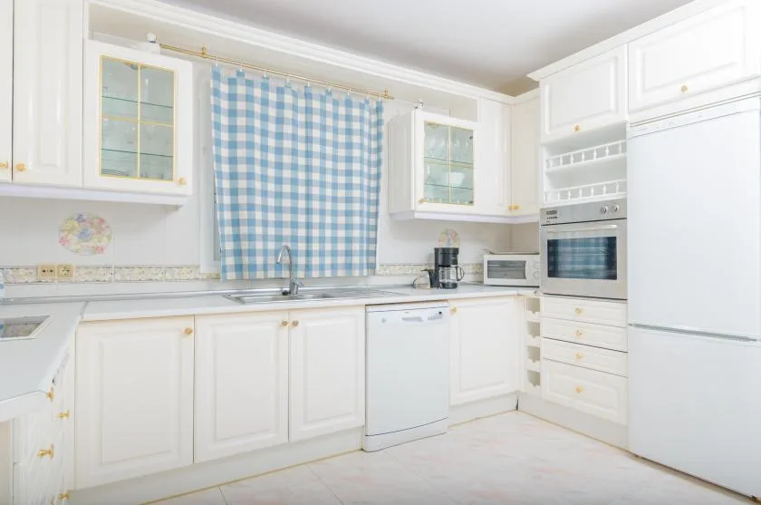 A bright white kitchen with a blue-and-white checked curtain above the sink.