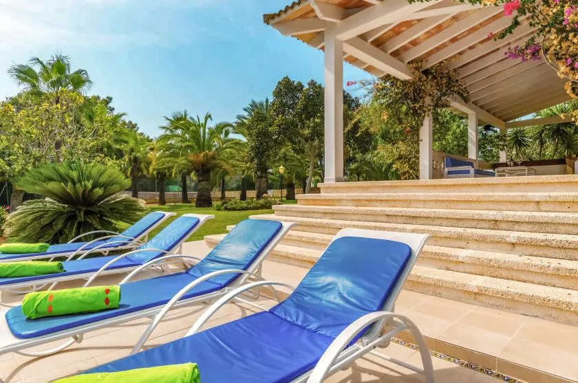 A poolside patio has blue loungers beside broad steps and a covered seating area.