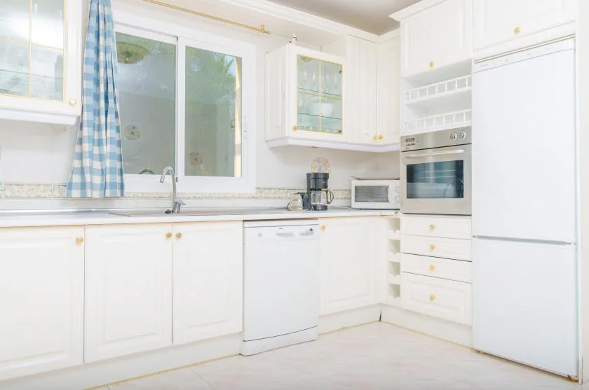 A bright white kitchen with a sink beneath the window and built-in appliances.