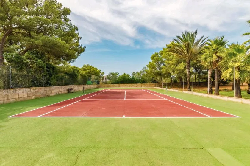 A red tennis court is surrounded by green grounds and trees.