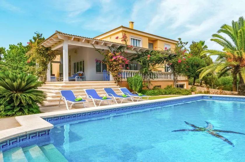 A house with a swimming pool and lounge chairs sits among lush garden plants.
