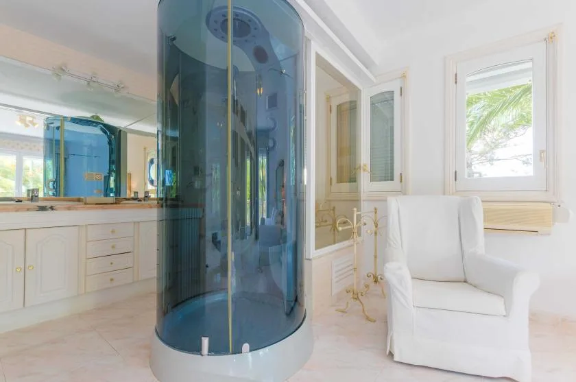 A bright bathroom with a blue curved shower enclosure and white seating.