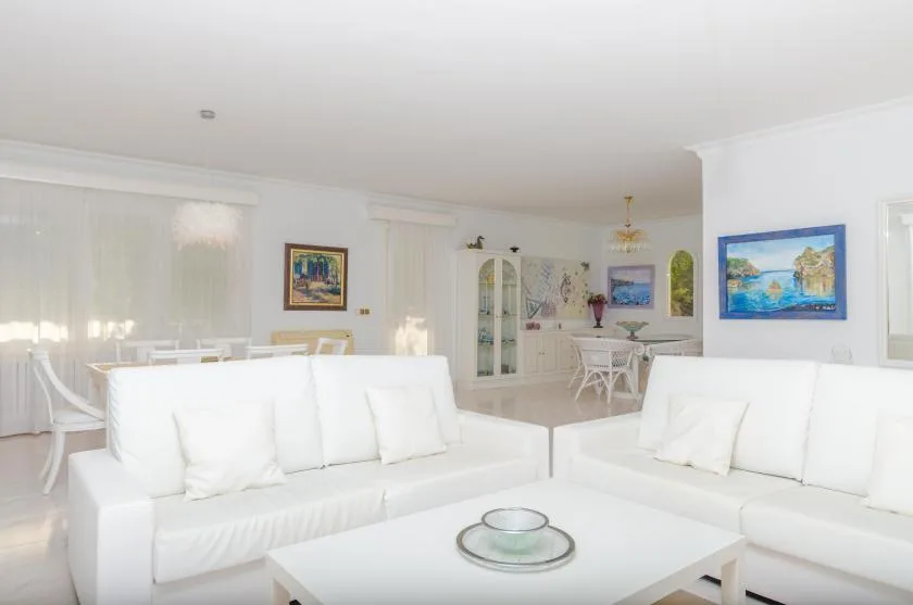 A bright living room with white seating and a dining area in the background.