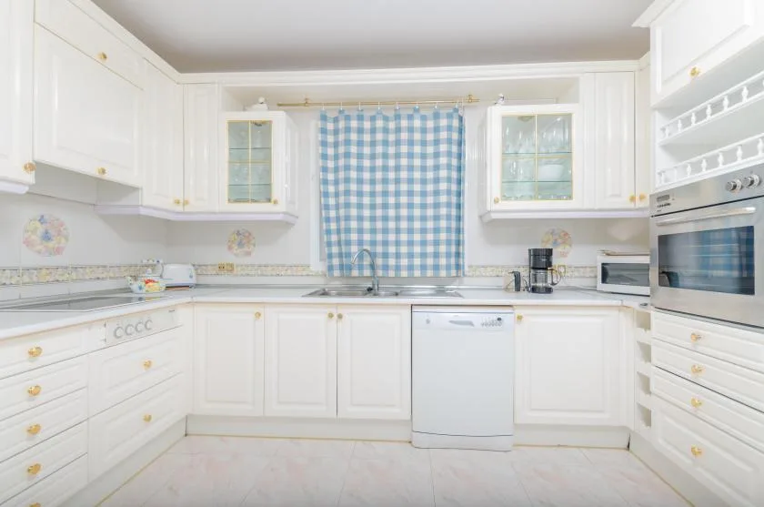 A bright white kitchen with a blue checkered curtain above the sink.
