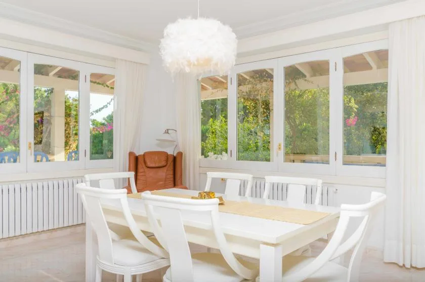 A bright dining room with a white table, seating, and large windows overlooking greenery.