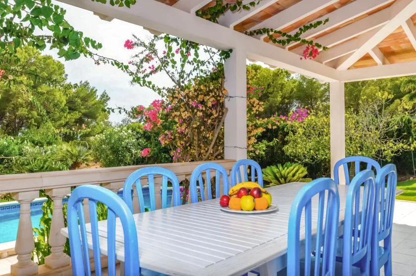 An outdoor dining area beneath a covered patio, surrounded by leafy plants.