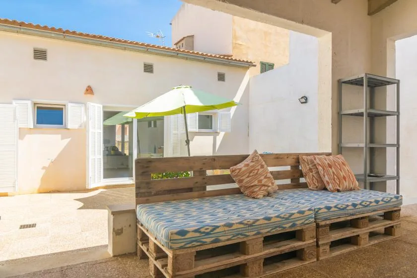 A sunlit patio features a cushioned wooden bench beneath a green-and-white umbrella.