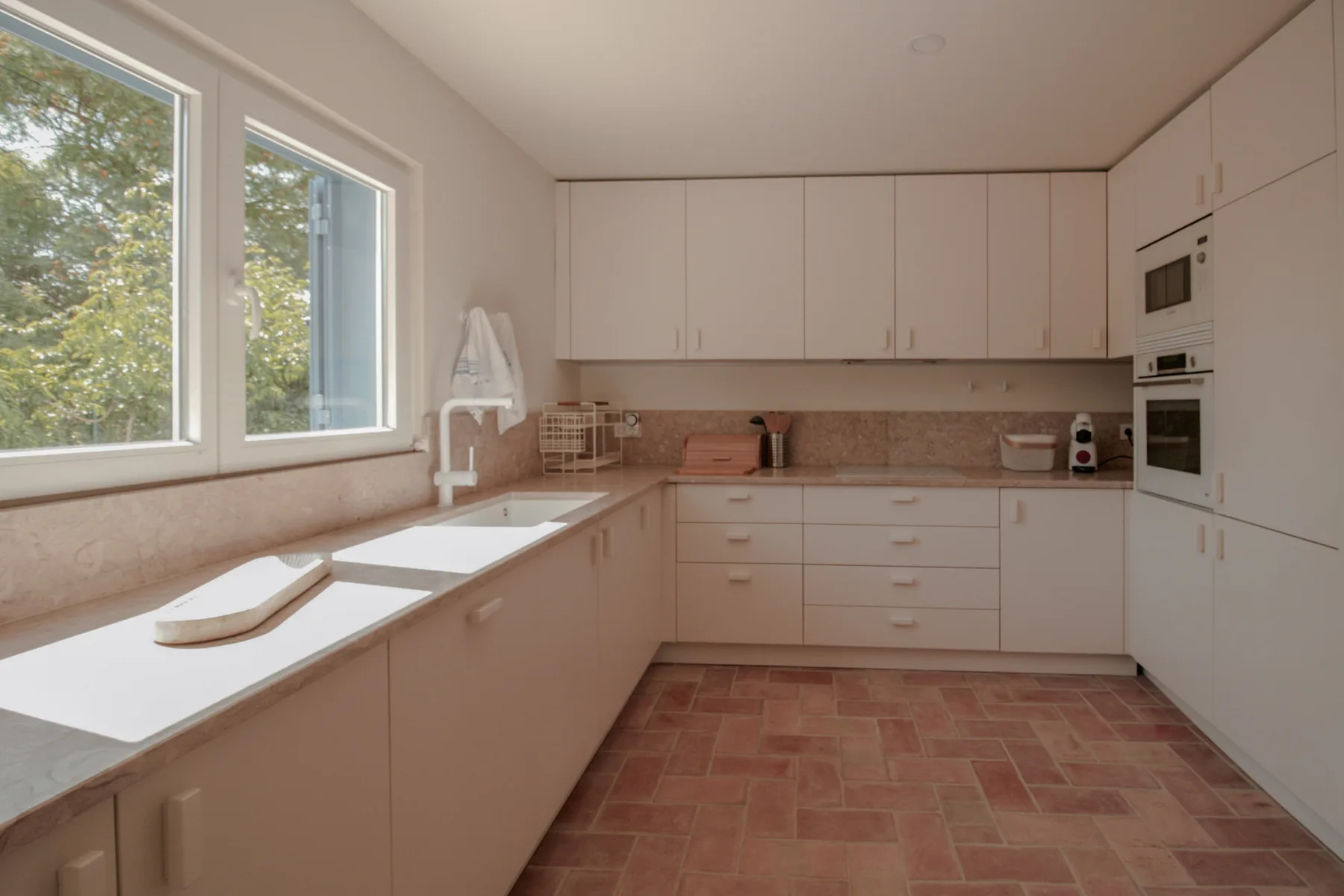 Una cocina luminosa con armarios blancos y una encimera larga bajo grandes ventanas.