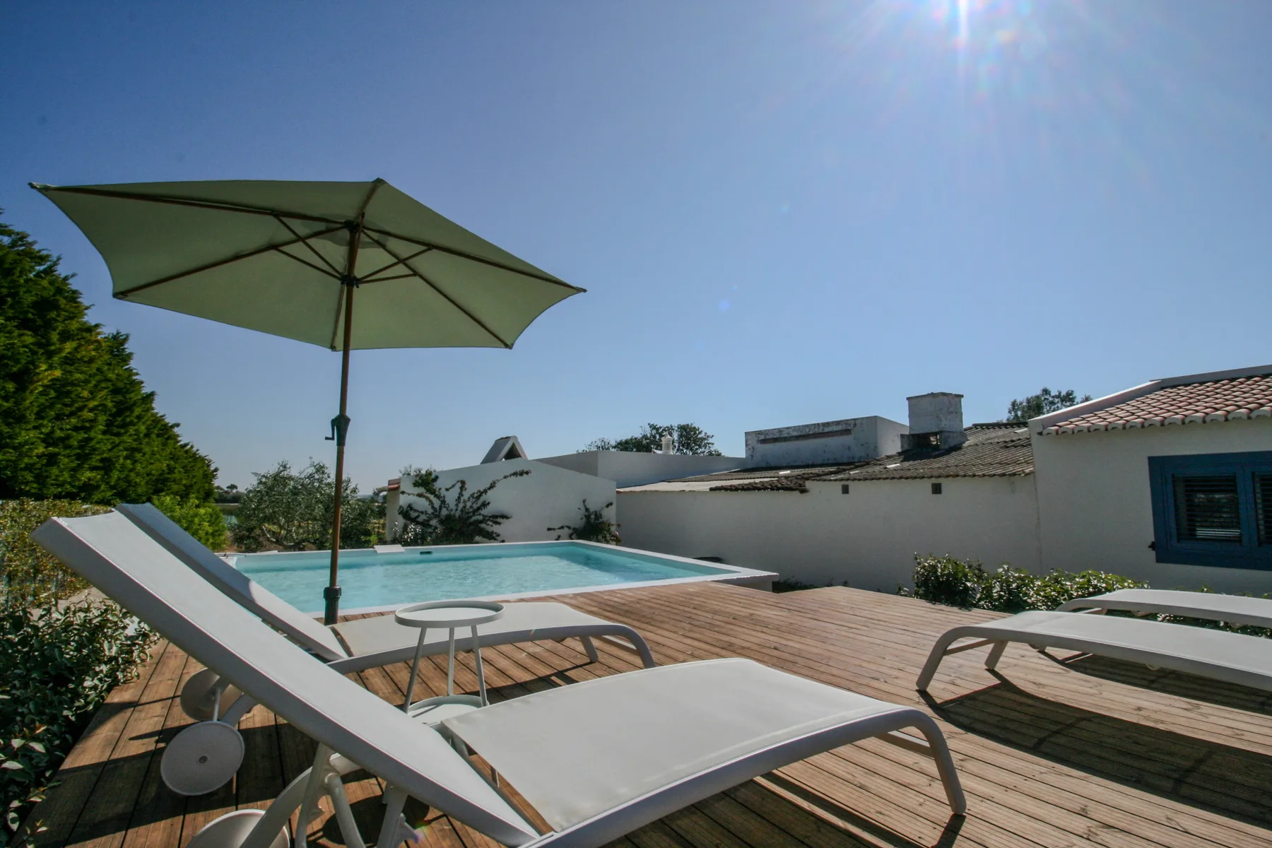 Una piscina al aire libre está junto a una terraza de madera con tumbonas y una sombrilla.