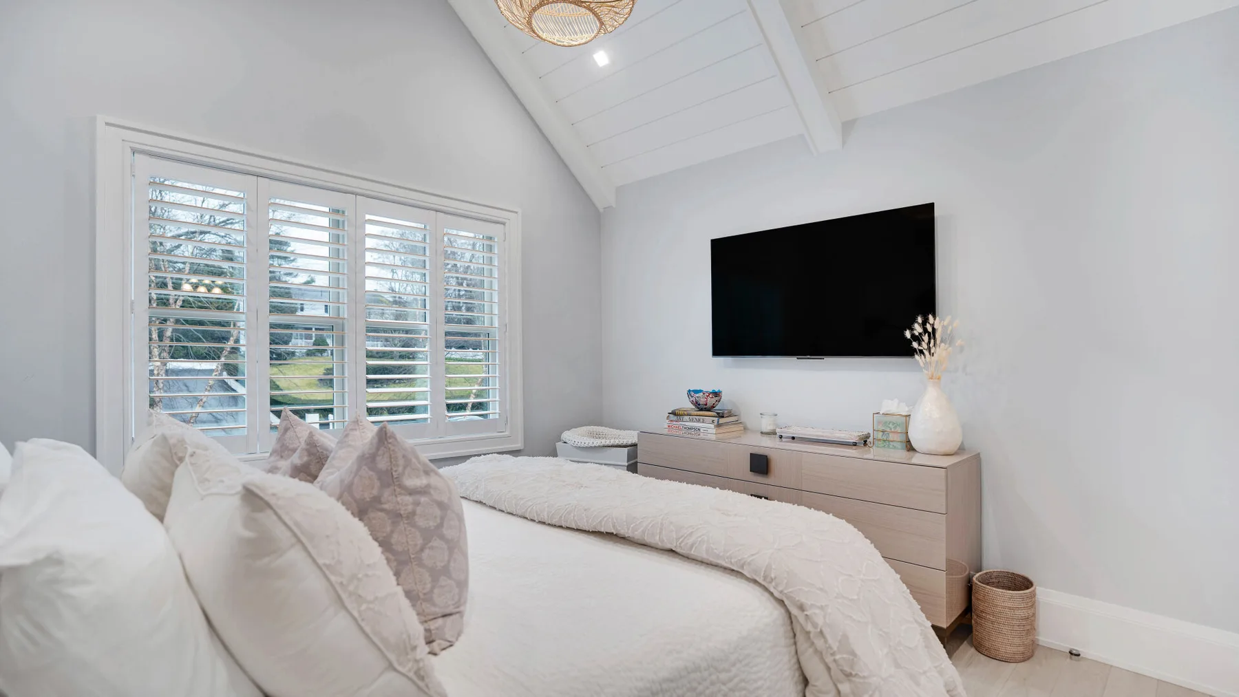 Un dormitorio luminoso con una cama, ventanas con persianas y un televisor en la pared.