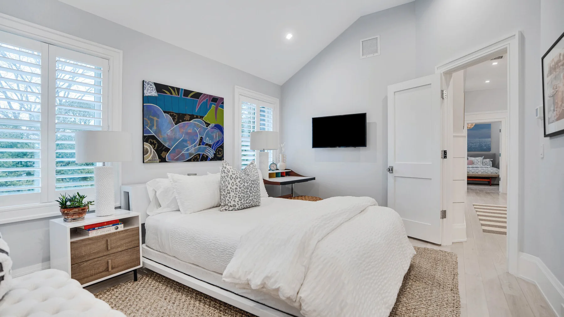Un dormitorio luminoso con una cama tendida y una obra de arte colorida en la pared.