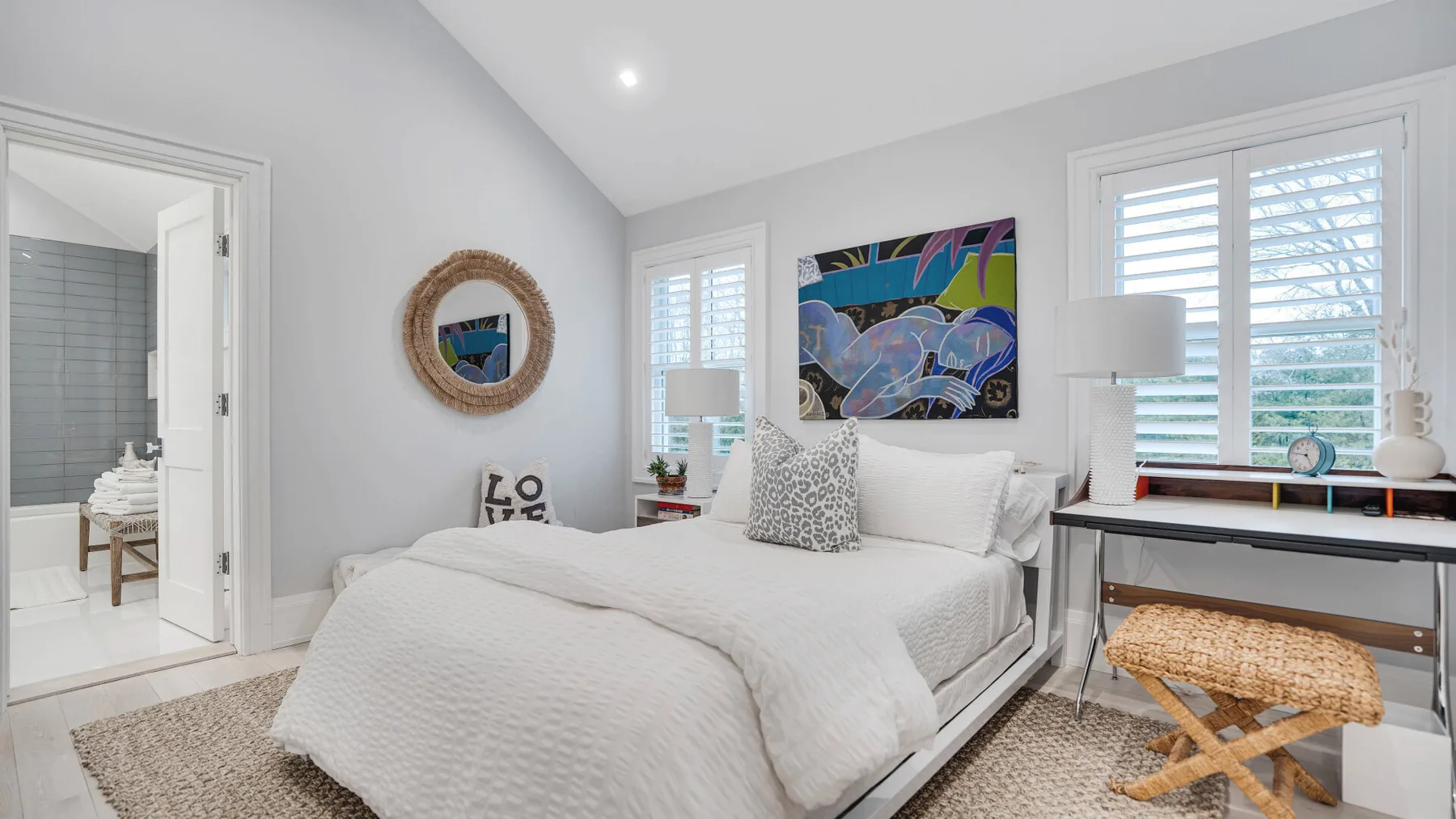 Un dormitorio luminoso con una cama tendida, arte colorido en la pared y ventanas amplias.