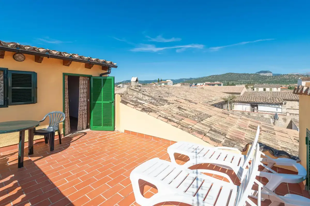 A rooftop terrace with white seating overlooks tiled rooftops and distant hills.
