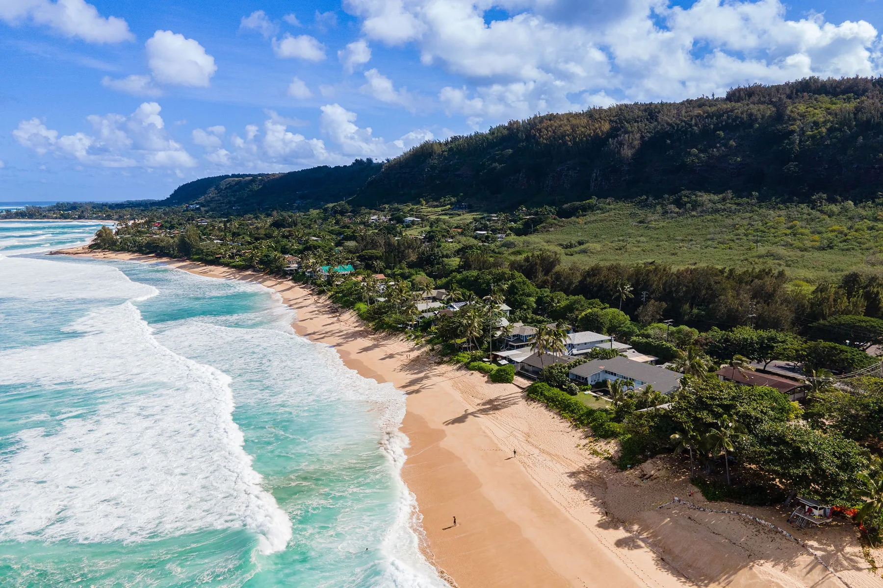 Vista aérea de una playa de arena junto a aguas turquesas y una ladera costera verde.