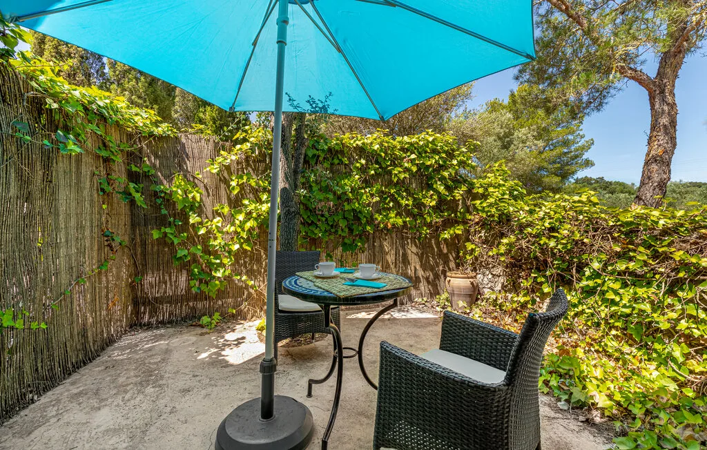 An outdoor patio with seating and a turquoise umbrella beside leafy greenery.