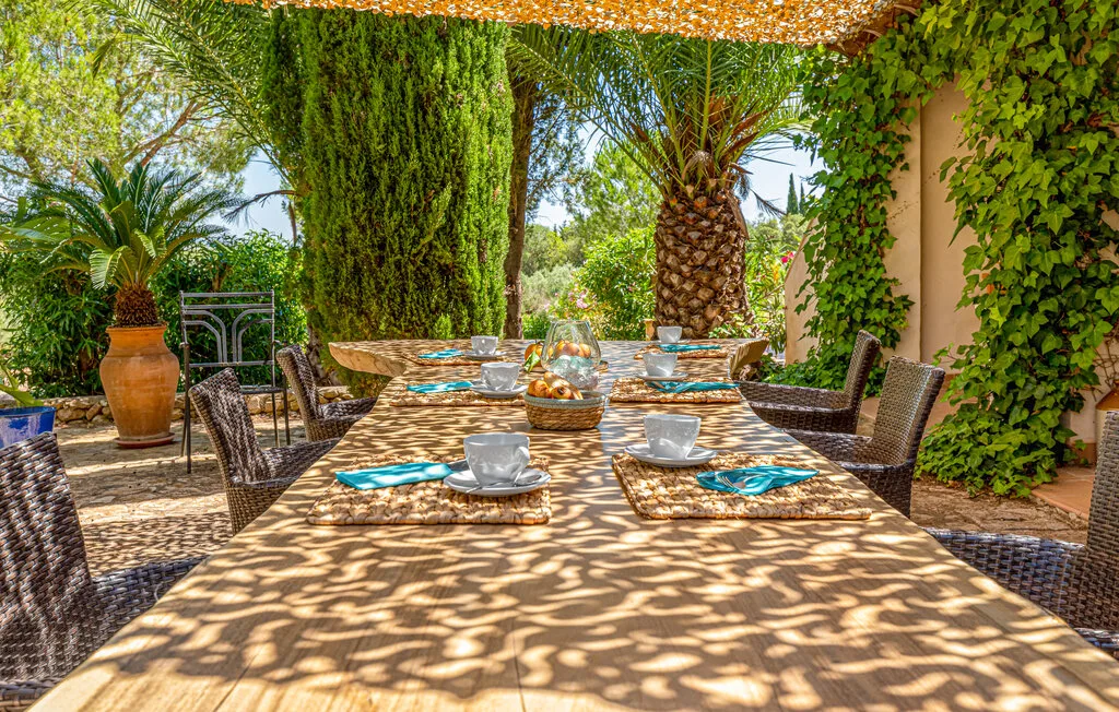An outdoor dining area with a long table set beneath a leafy pergola.