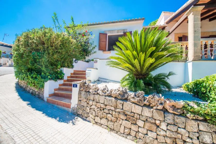 Entrada de una casa blanca con escalones, un muro de piedra y plantas.