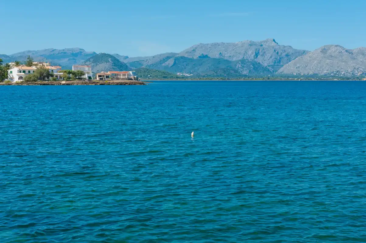 Una bahía costera con agua azul, casas en la orilla y montañas al fondo.