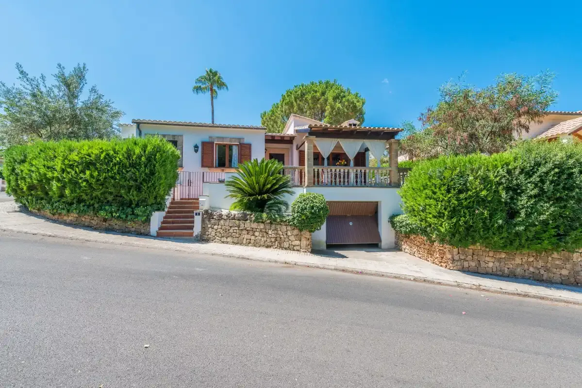 Una casa da a una calle pavimentada, con setos verdes y una palmera.