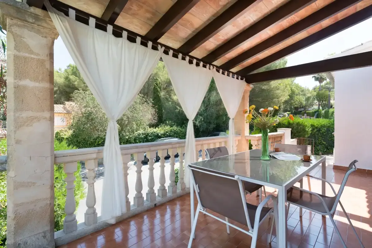 Un patio cubierto con una mesa de vidrio y cortinas blancas da a un jardín.