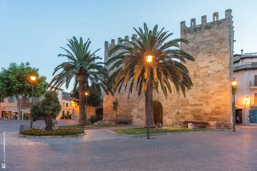 Una plaza pavimentada con palmeras se encuentra frente a un alto muro de piedra y una torre.