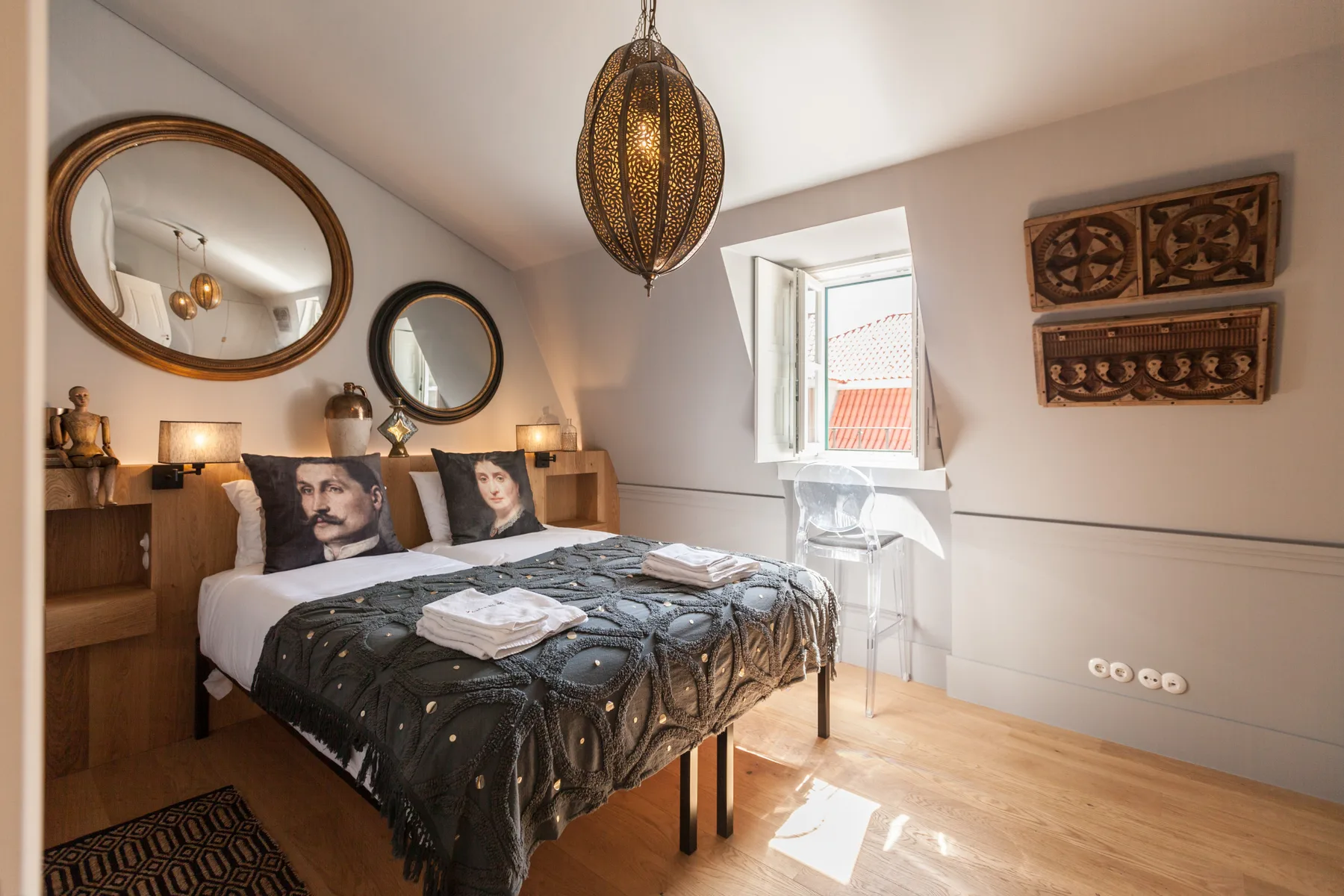 A bedroom with a bed dressed in dark patterned bedding beside an open window.