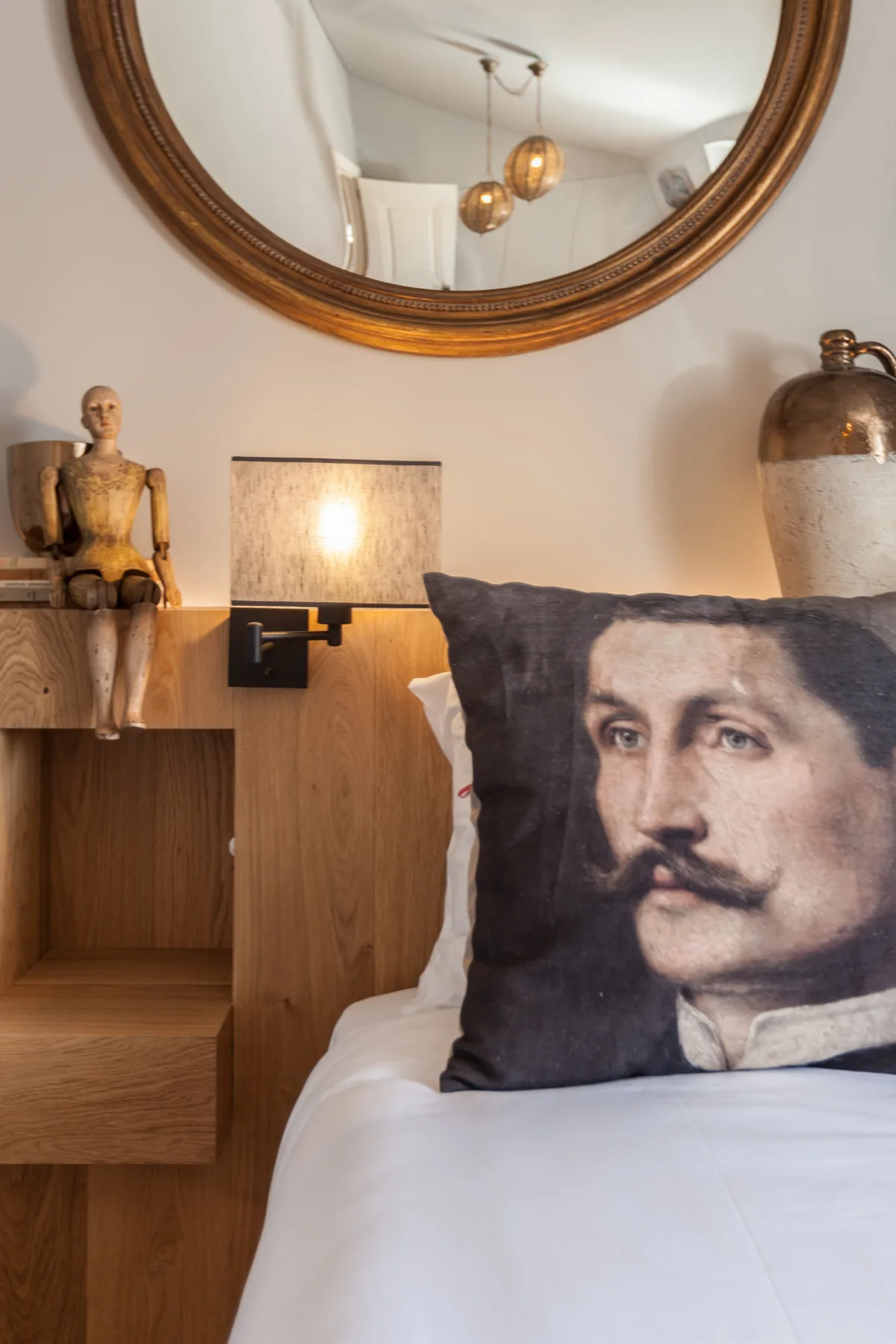 Dormitorio con una cama blanca, un cojín con un retrato y un espejo redondo en la pared.