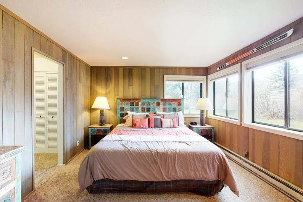 Un dormitorio con una cama entre dos mesitas y grandes ventanas.